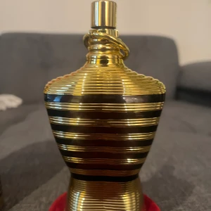 Jean Paul Gaultier Le Male Elixir - Jean Paul Gaultier Le Male Elixir parfym i en ikonisk guldig flaska formad som en manlig torso med svarta ränder. Kommer med en matchande guldig metallburk och röd sammetsbas. Flaskan har en lyxig och modern design som verkligen sticker ut.