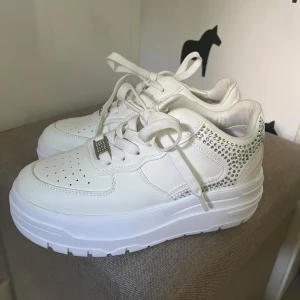 Vita chunky sneakers med strassdetaljer - Snygga vita chunky sneakers med platåsula och glittrande strassdetaljer på hälen. Skorna har snörning, perforerad tå och en dekorativ metallbricka med stenar på snörena. Bara provade, säljes pga fel storlek. Strl 33. 