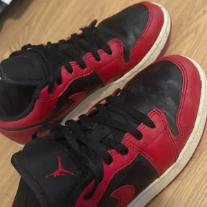 Nike Air Jordan 1 Low svart/röd - Säljer ett par Nike Air Jordan 1 Low i klassisk svart och röd färg. Skorna har svart ovandel i skinn med röda detaljer och vit mellansula. Röd yttersula och svarta skosnören. Ikonisk Jumpman-logga på plösen och Air Jordan-märke på hälen. Perfekta för dig som gillar streetwear.