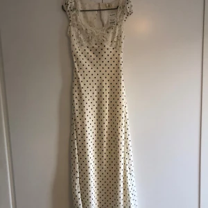 Vit långklänning med spets och prickar (polkadot) - Supersöt långklänning i vit satinlook med svarta prickar. Överdelen har vacker spets och små ärmar, samt en fin urringning. Klänningen har en feminin och romantisk vibe, perfekt för dig som gillar vintageinspirerat mode.