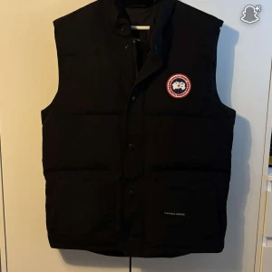 Svart väst från Canada Goose - Svart väst från Canada Goose med klassisk logga på bröstet. Västen är quiltad och har hög krage, två stora fickor framtill och knäppning med både dragkedja och tryckknappar. Perfekt för lager-på-lager under kyliga dagar.