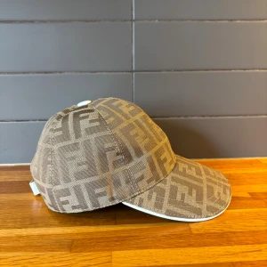 Beige Fendi keps med FF-mönster - Snygg beige keps från Fendi med det ikoniska FF-logomönstret över hela kepsen. Justerbar rem baktill i ljust läder med Fendi-logga. Kepsen har böjd skärm med vit kant och är tillverkad i ett slitstarkt tygmaterial. Perfekt för dig som vill sticka ut med en lyxig accessoar.