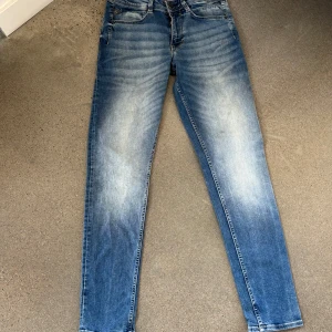 Rocko Slim Fit jeans blå W30 L32 - Snygga blå rocko jeans från garcia i modellen Slim Fit med smal midja och avsmalnande ben. Jeansen har klassisk femficksdesign och snygga slitningar framtill för en cool look. Perfekta till vardags med en hoodie eller t-shirt.