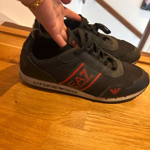 Svarta EA7 Emporio Armani sneakers - Snygga svarta sneakers från EA7 Emporio Armani med röda detaljer och logga på sidan samt sula. Skorna har snörning och är tillverkade i en mix av syntet och textil för en sportig look. Perfekta för dig som gillar stilrena och trendiga sneakers.