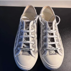 Christian Dior gråvita sneakers - Snygga sneakers från Christian Dior i grått och vitt med monogrammönster och tydlig logga på snörningen. Skorna har rund tå, platt sula och är tillverkade i textil med gummidetaljer. Perfekta för dig som vill ha en lyxig och trendig look.
