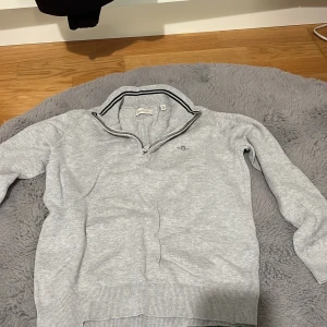 Grå half zip tröja från Gant - Ljusgrå tröja från Gant med half zip och ståkrage med svart-vit randig detalj. Broderad Gant-logga på bröstet. Tröjan har långa ärmar och ribbade muddar vid ärmslut och nederkant. Perfekt för en clean och sportig look.