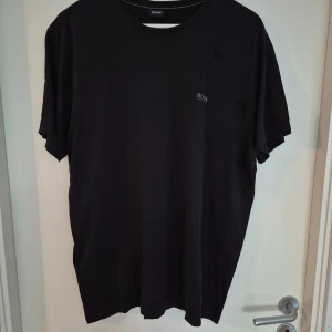 Svart t-shirt från Hugo Boss - Svart t-shirt från Hugo Boss med diskret logga på bröstet och större logga på axeln. Klassisk rund halsringning och korta ärmar. Tillverkad i mjuk bomull för en skön känsla och enkel stil.