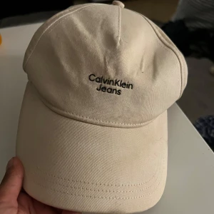 Beige keps från Calvin Klein Jeans - Snygg beige keps från Calvin Klein Jeans med broderad svart logga framtill. Klassisk böjd skärm och justerbar passform. Tillverkad i bomull för en skön och stilren look. Perfekt accessoar för att lyfta din outfit.