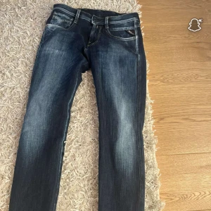 Replay blå jeans straight fit - Snygga blå jeans från Replay med klassisk straight fit och coola slitningar. Jeansen har fem fickor, ljusare tvättade partier och kontrastsömmar. Märkespatch baktill och Replay-logga på fickan fram. Perfekta för dig som gillar stilrena och tidlösa jeans.