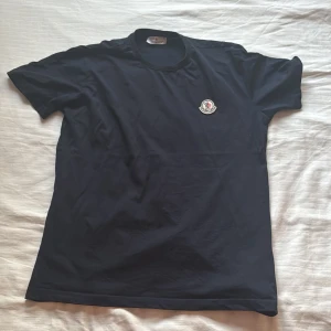 Svart Moncler t-shirt med logga - Svart t-shirt från Moncler i mjuk bomull. Klassisk rund hals och korta ärmar. På bröstet sitter Monclers ikoniska broderade logga i vitt, blått och rött. Perfekt basic med exklusiv känsla och snygg passform.