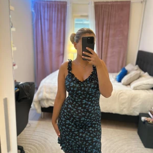 Svart blommig volangklänning - Supersöt svart kortklänning med blått blommönster och volangdetaljer vid axelband och nederkant. Klänningen har en figurnära passform och är ärmlös med v-ringning. Perfekt för dig som gillar romantisk och trendig stil.