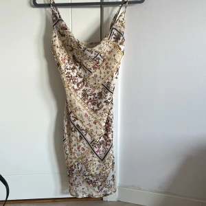 Blommig satinklänning med smala axelband - Säljer en superfin satinklänning med blommigt mönster i beige, brunt och inslag av rosa och gult. Klänningen har smala axelband, cowlneck och är figurnära med kort längd. Perfekt för sommaren och har en lyxig känsla tack vare det glansiga materialet.