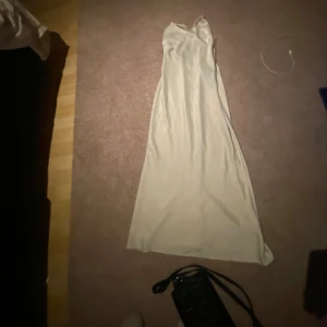 Lång ljus klänning från Nasty Gal - Supersnygg långklänning från Nasty Gal i en ljus, krämvit nyans. Klänningen har smala axelband, v-ringning och är figurnära med ett elegant fall. Materialet är glansigt och känns lyxigt, perfekt för speciella tillfällen.skicket är som nytt använt 1 gång och kommer inte till användning så de är darför jag säljer den 