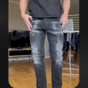 Svarta Dsquared2 jeans med färgstänk stl 48 - Säljer ett par svarta jeans från Dsquared2 med coola slitningar och färgstänk i rött och vitt. Jeansen har en straight fit och klassisk femficksdesign. Snyggt tvättade detaljer och diskret logga på baksidan. Lappar osv sitter kvar, passar dig som är ca 175-185