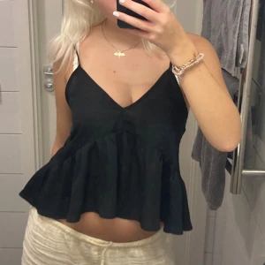 Svart volangtop från Gina Tricot - Supersöt svart linnetopp från Gina Tricot med volang nertill och smala justerbara axelband. Toppen har v-ringning och är luftig, perfekt till varma dagar eller att styla lager på lager. Materialet känns lätt och flowy.