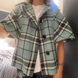 Rutig grön kappa med stora knappar - Säljer en kort, rutig kappa i grönt, svart och vitt med stora svarta knappar framtill. Jackan har bred krage, korta ärmar och en loose passform. Perfekt för dig som vill sticka ut med en cool och trendig look.
