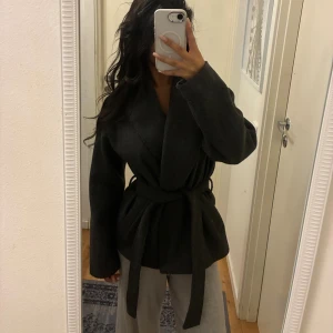 Mörkgrå omlottkappa från ONLY - Snygg mörkgrå omlottjacka från ONLY med bred krage och knytbälte i midjan. Jackan har lång ärm och en loose passform som ger en chill vibe. Materialet är ullblandning som känns mjukt och håller dig varm under höst och vinter. Köpt för 799kr