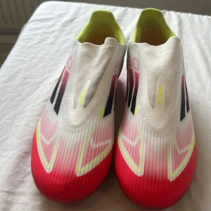 Adidas F50 fotbollsskor vit/röd - Säljer ett par Adidas F50 fotbollsskor i vitt, rött och neongult. Skorna har en strömlinjeformad design utan snörning, med svarta och röda detaljer på sidorna och neongul insida. Ovandelen är i syntetmaterial för lätt vikt och bra bollkontroll. Använda endast 2 gånger