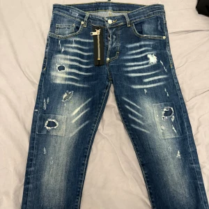 Blå slitna jeans med dragkedjedetalj - Säljer ett par blå jeans med coola slitningar och patchade hål på benen. Jeansen har en unik dragkedjedetalj vid fickan och snygga ljusa tvättade partier. Perfekt för dig som gillar streetstyle och vill sticka ut med din outfit.