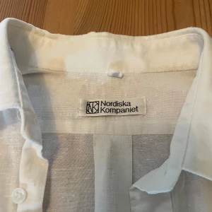 Vit linneskjorta Nordiska Kompaniet - Vit skjorta i linne från Nordiska Kompaniet med klassisk krage, korta ärmar och bröstficka med knapp. Skjortan har en avslappnad passform och knäppning framtill. Perfekt för varma dagar och enkel att styla med jeans eller shorts.