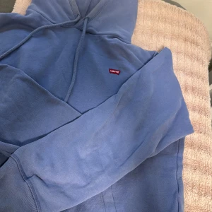 Blå hoodie från Levi's XS - Säljer en blå hoodie från Levi's i storlek XS. Tröjan har huva med dragsko, magficka och klassisk röd Levi's-logga på bröstet. Mjuk och skön bomullskvalitet, perfekt för chill dagar. Långärmad och med ribbade muddar vid ärmslut och nederkant.