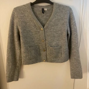 Grå kofta från H&M  - Mysig grå kofta från H&M med v-ringning och guldknappar framtill. Tröjan har två ribbade fickor och ribbade muddar vid ärmslut och nederkant. 