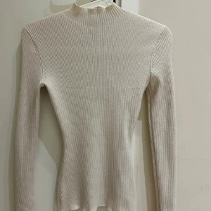 Beige ribbad polotröja - Säljer en beige ribbad polotröja med lång ärm och hög hals. Tröjan är figurnära och har en stilren, enkel design som passar perfekt till lager-på-lager. Materialet är mjukt och stretchigt för en bekväm känsla.