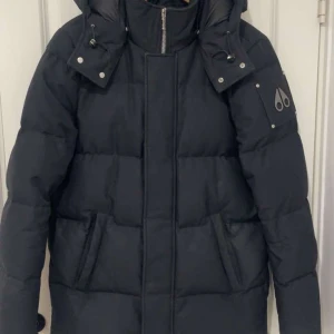 Svart dunjacka moose knuckles - Svart dunjacka från Moose Knuckles med huva och dragkedja framtill. Jackan har quiltad design, två stora fickor och logotyp på ärmen. Perfekt för kalla dagar med sin tjocka och varma känsla. Snygg och stilren modell som håller dig varm hela vintern. Jackan har använts i 2 vinter och har inga skador. Jackan ligger på 13-15 tusen och köpt för 11 tusen.