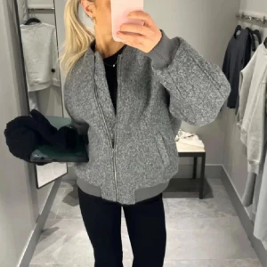 Grå bomberjacka i ullmix - Säljer en grå bomberjacka med oversized passform och ribbade muddar. Jackan har dragkedja framtill och är tillverkad i en ullblandning som ger en mjuk och lite fluffig känsla. Perfekt för dig som gillar en chill och trendig look.