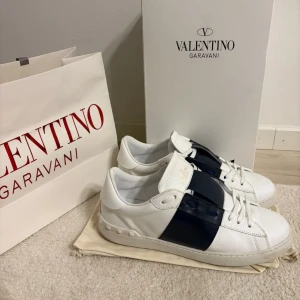 Valentino Garavani Open sneakers  - Snygga Valentino Garavani Open sneakers i vitt skinn med bred marinblå rem över vristen. Klassisk rund tå, snörning och ikoniska nitar baktill på sulan. Lyxig och stilren design, perfekt för dig som gillar exklusiva sneakers.