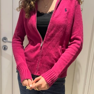 Mörkrosa kofta från Ralph Lauren Sport - Mörkrosa kofta från Ralph Lauren Sport i 100% bomull. Dragkedja framtill, ribbade muddar och fickor på sidorna. Klassisk broderad logga på bröstet och långärmad modell. Perfekt för lager-på-lager och snygg till jeans.