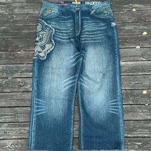 Blå wide jeans från Akademiks - Riktigt feta blå jeans från Akademiks med bred passform och stora broderade patches på både fram- och baksida. Snygga slitningar och coola detaljer som sticker ut. Perfekta om du vill ha en streetig vibe och unik look.