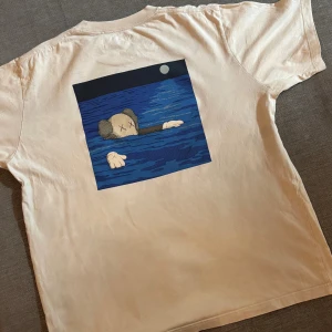 Vit KAWS x Uniqlo t-shirt med tryck - Säljer en beige t-shirt från Uniqlo i samarbete med KAWS. Framsidan har ett litet tryck av KAWS Companion som simmar, och baksidan har en stor grafisk illustration i blått och svart med måne. Klassisk passform och rund hals, tillverkad i mjuk bomull. Storlek L