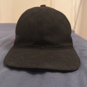 Svart keps från Gucci Canvas - Snygg svart keps från Gucci med klassisk röd och grön rand på sidan. Kepsen har böjd skärm, justerbart spänne bak i skinn och diskret GG-mönster i tyget. Perfekt accessoar för en trendig look.