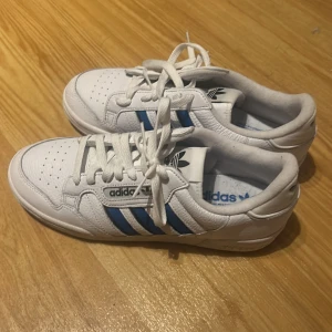 Adidas sneakers med blå ränder - Snygga vita Adidas sneakers med klassiska blå ränder på sidorna och svart Adidas-logga på tungan. Skorna har snörning, rund tå och är tillverkade i skinn med vit sula. Perfekta för dig som gillar retrostil och streetwear.