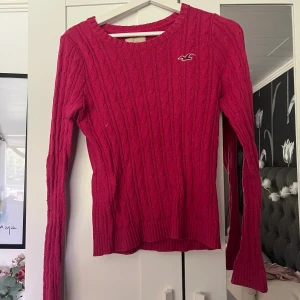 Rosa stickad tröja från Hollister - Säljer en snygg rosa stickad tröja från Hollister med kabelstickat mönster och rund hals. Tröjan har lång ärm och en liten vit Hollister-logga på bröstet. Perfekt för dig som gillar klassisk och enkel stil med en poppig färg. 