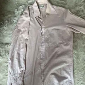 Ljusrosa skjorta från Melka - Säljer en stilren ljusrosa skjorta från Melka. Skjortan har klassisk krage, bröstficka och knäppning framtill. Tillverkad i mjuk bomull och har långa ärmar med manschettknappar. Perfekt för dig som vill ha en clean och enkel look.