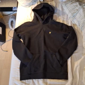 Svart hoodie från Lyle & Scott Junior - Svart hoodie från Lyle & Scott Junior med klassisk gul logga på bröstet. Tröjan är en varm och skön tröja som är bra till kallare dagar och svala kvällar. Om ni har frågor är det bara att skriva☺️