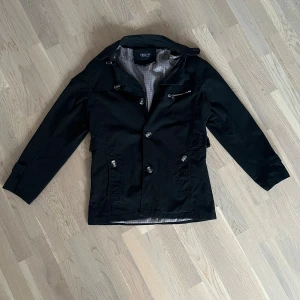 Svart rock från Fashion med detaljer - Svart rock med knappar och silver dragkedja på bröstet. Jackan har klassisk krage, axelklaffar och justerbara remmar i midjan. Tillverkad i ett slätt material som känns lätt men ändå robust. Perfekt för dig som gillar stilrena och snygga ytterplagg.