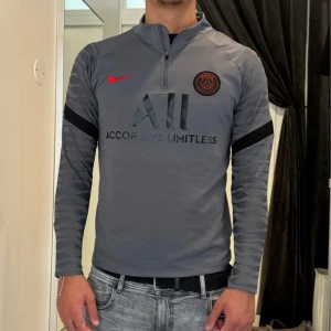 Grå PSG långärmad träningströja Nike - Säljer en grå långärmad Paris Saint-Germain träningströja från Nike med svart och rött PSG-emblem, röd Nike-logga och svart tryck. Ärmarna har subtilt mönster och svart band. Halv dragkedja framtill och tryck på ryggen. Perfekt för fotboll eller streetwear.
