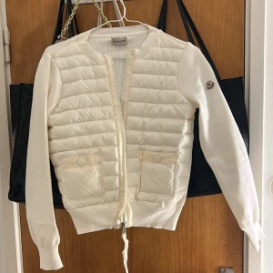 Vit cardigan jacka från Moncler - Snygg vit cardigan jacka från Moncler med två stora fickor framtill och dragkedja. Jackan har långärmade stickade ärmar och vadderad front. Klassisk Moncler-logga på ena ärmen. Perfekt för dig som vill ha en stilren och exklusiv look.
