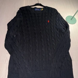 Svart kabelstickad tröja Polo Ralph Lauren - Svart kabelstickad tröja från Polo Ralph Lauren med klassisk röd broderad logga på bröstet. Rund hals och ribbade muddar vid ärmslut och nederkant. Perfekt för dig som gillar stilrena och tidlösa plagg med premiumkänsla.