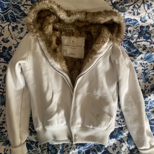 Vit hoodiejacka med fuskpäls från Abercrombie - Vit hoodiejacka från Abercrombie & Fitch med mysig brun fuskpäls i huvan. Liknar jackan jennifer check har på sig i jennifers body. Assnygg jacka som funkar perfekt till hösten och våren!! Den har en liten fläck, och lite missfärgning på insidan av jackans elastiska band, den är även lite nopprig vid de elastiska banden! Jag har även inte gjort några försök att ta bort fläcken elller missfärgningen! Står storlek S på lappen, men passar nog även M, jag bär storlek XS som referens 💕