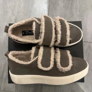 Grå sneakers med fuskpäls från Duffy - Mysiga grå sneakers från Duffy med breda kardborreband och fluffig beige fuskpäls runt kanterna och inuti skon. Ovandelen är i mockaliknande material och sulan är vit och platt. Perfekta för dig som vill ha både stil och komfort. ENDAST ANVÄNDA TVÅ GÅNGER ❤️