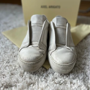 Vita skor från Axel Arigato - Säljer ett par stiliga vita sneakers från Axel Arigato. Skorna är i skinn med minimalistisk design, platt sula och rund tå. Diskret logga i guld på sidan och elastisk panel framtill istället för klassisk snörning. Perfekta för en clean och modern look.