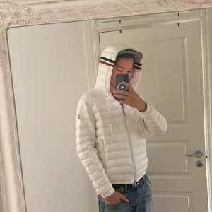 Vit Moncler dunjacka med huva - Säljer en snygg vit dunjacka från Moncler med huva och coola ränder i rött och marinblått på kanten. Jackan har dragkedja framtill, två sidofickor med dragkedja och justerbar snörning vid huvan. Perfekt för dig som vill ha en clean och stilren look. Storlek 2/M