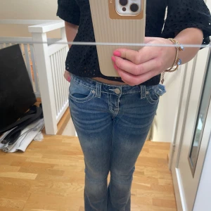 True Religion blå bootcut jeans - Snygga blå jeans från True Religion med låg midja och bootcut passform. Jeansen har tydliga vita kontrastsömmar, klassiska fickor fram och bak samt dekorativa broderier på bakfickorna. Tillverkade i stretchig denim för en bekväm känsla. Har klippt dem där nere för dem var för långa