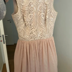 Champagnefärgad festklänning  - Säljer en elegant kortklänning i champagnefärg med glittriga paljettdetaljer och broderat mönster på överdelen. Kjolen är mjukt veckad och ger en flowy känsla. Klänningen är ärmlös och har en v-ringad rygg med dragkedja. Perfekt för dig som vill sticka ut med stil.          Passar bäst för 34/ XS