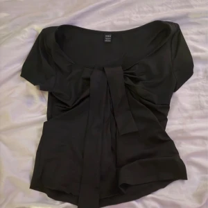 Svart topp med knyt och djup urringning - Svart topp från SHEIN med korta ärmar, djup urringning och snygg knytning framtill. Modellen är figurnära och har en croppad känsla. Perfekt att styla med jeans eller kjol för en trendig look. Aldrig använtd