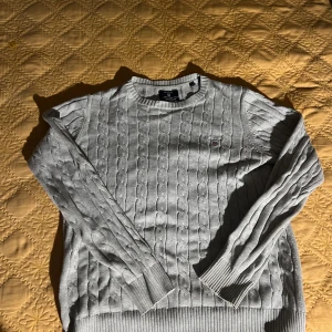 Ljusgrå kabelstickad tröja från Gant - Säljer en ljusgrå kabelstickad tröja från Gant i premium bomull. Tröjan har rund hals, ribbade muddar och diskret Gant-logo på bröstet. Perfekt för lager-på-lager och passar till både jeans och chinos.